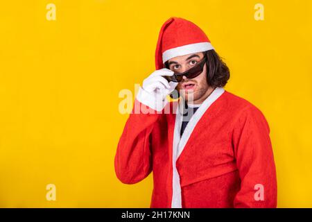 Un giovane Santa sorpreso! Un bell'uomo vestito come Babbo Natale si toglie gli occhiali da sole con un'espressione sorpresa sul viso. Sfondo giallo. Foto Stock