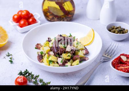 Insalata calda con polpo, patate, pomodori, capperi e limone su un piatto bianco. Primo piano, sfondo bianco. Foto Stock