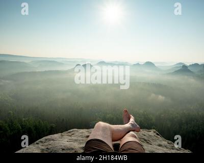 Uomo viaggiatore e turista hanno un riposo sulla cima alta montagna. Gambe maschi incrociate. Vista soleggiata di mattina presto nel paesaggio sognante. Foto Stock