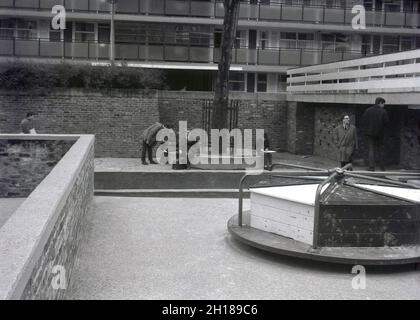 1960, storico, un parco giochi in cemento e murato, in un grande complesso residenziale del consiglio, Churchill Gardens a Pimlico, Inner London, Inghilterra, Regno Unito. Costruito tra il 1948 e il 1962 per sostituire le terrazze vittoriane danneggiate nel Blitz della seconda Guerra Mondiale, questo era un enorme patrimonio di alloggi sociali, con 32 blocchi torre che forniscono 1,600 case. All'esterno degli appartamenti, nel recinto murato parco giochi con una base in cemento, una tradizionale rotonda in legno e metallo. Foto Stock