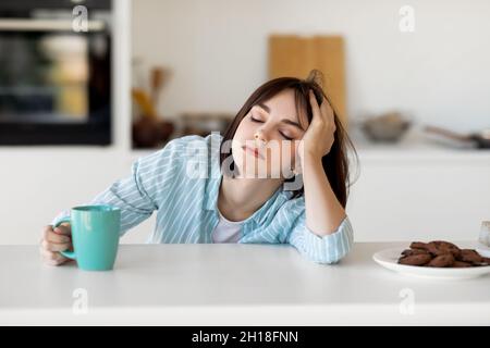 Donna addormentata che beve caffè, si sente stanca, soffre di insonnia e disturbi del sonno, seduta in cucina Foto Stock