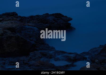 LUNGA ESPOSIZIONE NOTTURNA, IMMAGINE DEL CIELO DRAMTIC E ONDA DI MARE CON ROCCIA IN SFONDO SCENARIO. Foto Stock