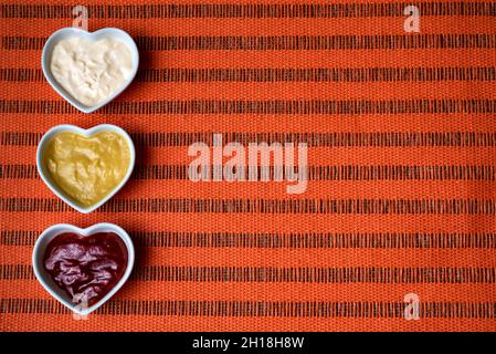 Ciotole a forma di cuore con ketchup, maionese e salsa di senape isolate su un lato su uno sfondo arancione ruvido.Copy-space. Foto Stock