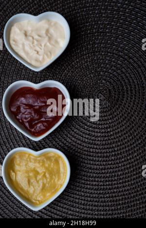 Ciotole a forma di cuore con ketchup, maionese e salsa di senape isolate su un lato su uno sfondo marrone.Copy-sapce verticale. Foto Stock