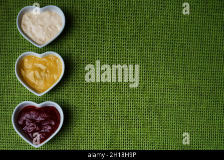 Piccole ciotole a forma di cuore con ketchup, maionese e salsa di senape isolate su un lato su un tessuto verde ruvido background.Copy-space. Foto Stock