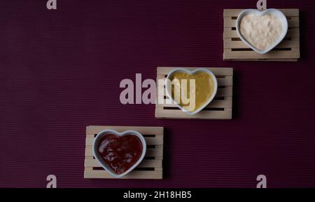 Piccole ciotole a forma di cuore su piccoli pallet con ketchup, maionese e salsa di senape che formano una scala su uno sfondo di maroon.Copy-space. Foto Stock