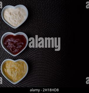Ciotole a forma di cuore con ketchup, maionese e salsa di senape isolate su un lato su uno sfondo marrone. Foto Stock