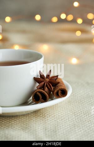 Tazza bianca di tè con cannella su quadri beige alle luci di Natale. Natale ancora vita. Foto Stock