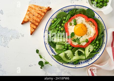 Peperoni rossi ripieni di uova, foglie di spinaci, piselli verdi e micrograni su un piatto per la colazione su sfondo grigio chiaro. Vista dall'alto. Foto Stock