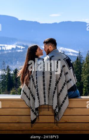 Coppia amorevole avvolta in una coperta seduta sulla veranda di una villa di montagna sullo sfondo dei Carpazi, vacanza in famiglia. Foto Stock