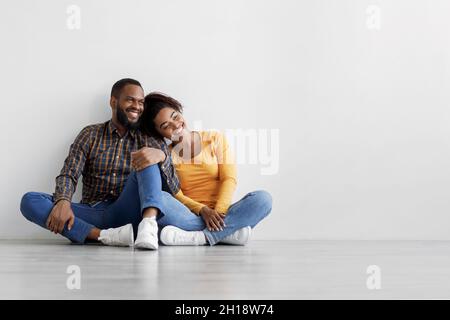 Soddisfatto giovane afroamericano maschio e femmina seduta sul pavimento su sfondo grigio parete, abbraccio, progettazione interni Foto Stock