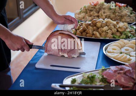 Mano dello chef maschile con il coltello di carne che affetta un giunto di maiale arrosto croccante su una tavola bianca posizionata su un tavolo con grandi piatti di patate insalate Foto Stock