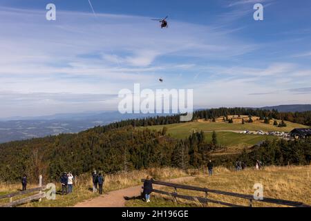 Waldkirch, Germania. 15 ottobre 2021. Gli spettatori si levano in piedi sulla cima del Kandel mentre sullo sfondo due persone appendono su una corda sotto un elicottero di salvataggio del Luftrettung DRF. Durante un allenamento con verricello, l'equipaggio della stazione di Friburgo della DRF Luftrettung (cartello di chiamata Christoph 54) si pratica con il verricello sotto un elicottero insieme al Bergwacht Schwarzwald. Questo verricello può essere utilizzato per salvare i pazienti da terreni impraticabili, nonché per trasportare medici e paramedici in regioni difficili da raggiungere. Credit: Rin/dpa/Alamy Live News Foto Stock