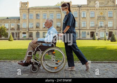 Scatto a tutta lunghezza di una giovane infermiera che indossa visiera e maschera che cammina con paziente uomo maturo in sedia a rotelle Foto Stock