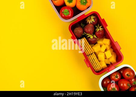 Scatole da pranzo con biscotti, pezzi di formaggio, fragole, pomodori e peperoni su sfondo giallo. Pasto scolastico e pranzo d'affari. Vista superiore Foto Stock