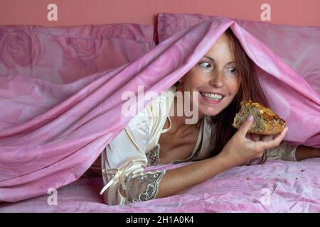 Notte tema gluttony, donna sul letto che mangia segretamente un pezzo di torta Foto Stock