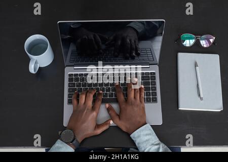 Vista dall'alto minima di un uomo afro-americano irriconoscibile che utilizza un computer portatile mentre lavora su sfondo nero della scrivania, concentrarsi sulle mani alla tastiera, spazio per la copia Foto Stock