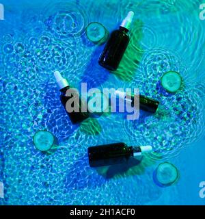 Bottiglie cosmetiche contagocce e tagliare il cetriolo in acqua su sfondo colorato Foto Stock