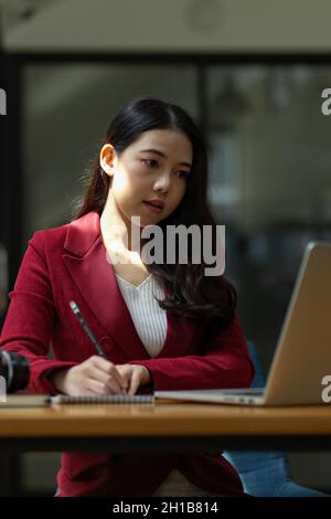 Donna d'affari giovane focalizzata usando il computer portatile e facendo qualche lavoro di ufficio o prendendo note di importanza sui notepad in ufficio. Foto Stock