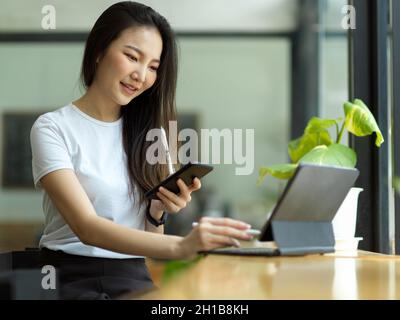 Bella ragazza asiatica freelance che usa smartphone e lavora su un tablet portatile al bar. Foto Stock