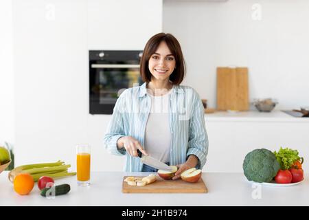 Felice signora giovane tagliare frutta su tavola di legno, preparare insalata fresca, in piedi in cucina e sorridendo alla macchina fotografica Foto Stock