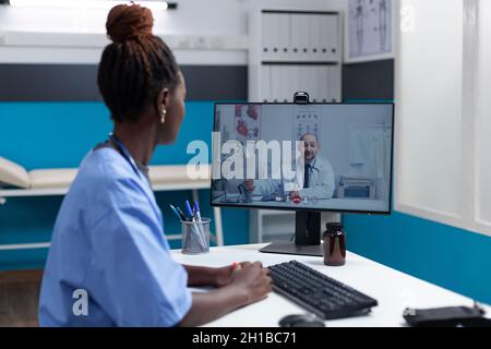 Infermiera afroamericana che spiega i sintomi della malattia al medico remoto in attesa di consiglio durante la conferenza online di videoconferenza che lavora in ospedale ufficio. Chiamata telefonica sullo schermo del computer Foto Stock