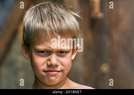 Scolboy junior arrabbiato con capelli biondi ruvidi in posa per la macchina fotografica nel cortile della casa d'estate su bella vista giorno d'estate vicino Foto Stock