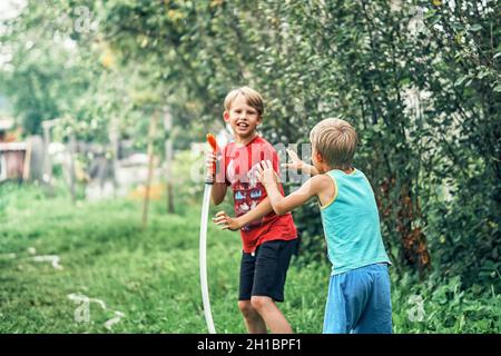 Gioiosa ragazzo divertente con simpatico fratellino gioca con il tubo d'acqua nel giardino verde in un ampio cortile estivo in bella giornata di sole Foto Stock