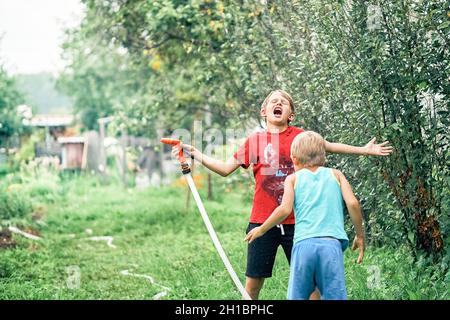 Gioiosa ragazzo divertente con simpatico fratellino gioca con il tubo d'acqua nel giardino verde in un ampio cortile estivo in bella giornata di sole Foto Stock