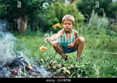 Ragazzo divertente arrostire un pezzo di pane sul bastone sopra falò seduto sul prato verde erba durante la vacanza in bella giornata estiva Foto Stock