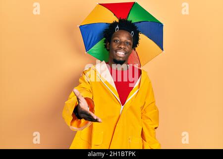 Giovane uomo afroamericano che indossa un impermeabile giallo che sorride allegro offrendo mano di palma che dà assistenza e accettazione. Foto Stock
