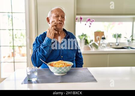 Uomo anziano con capelli grigi mangiare spaghetti di pasta a casa toccando la bocca con mano con espressione dolorosa a causa di mal di denti o malattie dentistiche su te Foto Stock