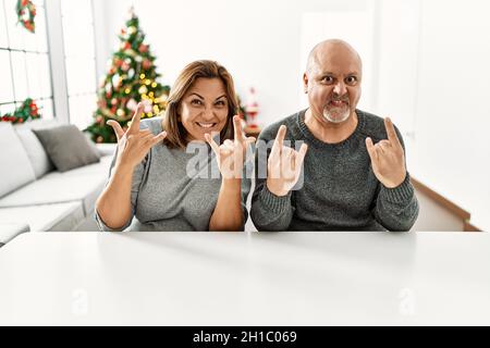 Coppia ispanica di mezza età seduta sul tavolo da albero di natale gridando con pazza espressione facendo rock simbolo con le mani in su. Star della musica. Heavy co Foto Stock