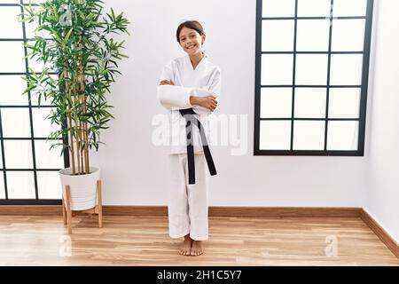 Ragazza ispanica giovane che indossa kimono karate e cintura nera faccia felice sorridente con le braccia incrociate che guardano la macchina fotografica. Persona positiva. Foto Stock