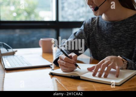 Primo piano mano di donna asiatica che studia online da casa facendo appunti per gli studenti imparare a distanza su laptop facendo i compiti, guardando il video di ascolto Foto Stock