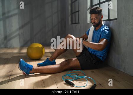 Giovane sportivo afroamericano seduto e utilizzando smartwatch al coperto in palestra, allenamento Foto Stock