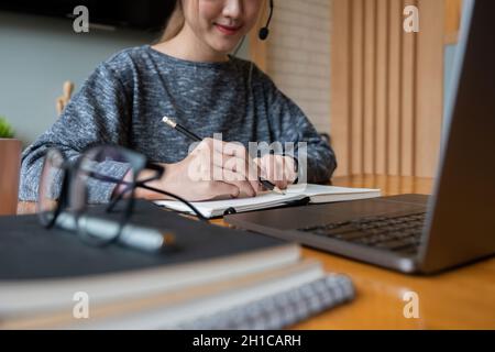 Primo piano mano di donna asiatica che studia online da casa facendo appunti per gli studenti imparare a distanza su laptop facendo i compiti, guardando il video di ascolto Foto Stock