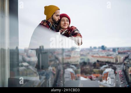 Giovane coppia avvolta in coperta in piedi e abbracciato all'aperto sul balcone, fucilato attraverso il vetro. Foto Stock
