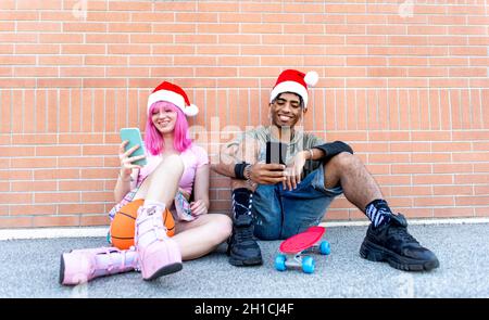 buon natale! felice coppia diversificata di amici che indossano natale santa cappello che interagiscono con smartphone seduta su parete di fondo insieme in inverno Foto Stock