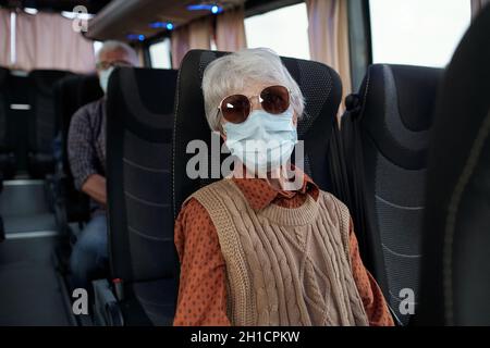 Donna matura con capelli grigi in maschera protettiva seduta sul sedile doppio all'interno dell'autobus durante il giro interurbano Foto Stock