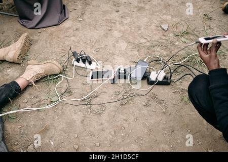 Mano di giovane rifugiato che tiene lo smartphone in carica mentre si siede accanto ad altri gadget, spine e caricabatterie Foto Stock