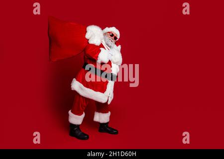 Foto del profilo a tutta lunghezza del peso di santa portare borsa grande con presenta cappello di abbigliamento giacca pantaloni stivali isolati su sfondo rosso Foto Stock