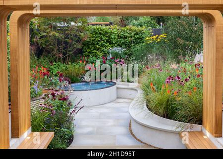Trovare il nostro modo: Un NHS Tribute Garden. Pergola in legno conduce in un cortile pavimentato sunken con pavimentazione bianca, piscina rialzata e aiuole fiorite. Piante Foto Stock