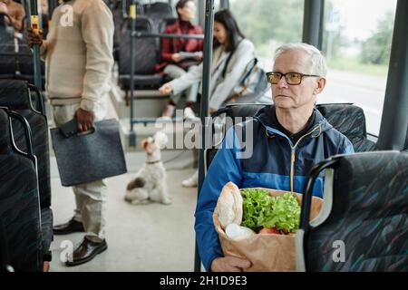 Ritratto di uomo anziano seduto in autobus mentre si viaggia con i mezzi pubblici in città e tenendo una borsa di carta con generi alimentari, spazio copia Foto Stock