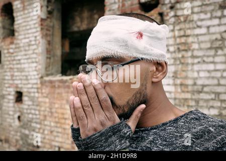 Giovane rifugiato maschio ferito con bendaggio intorno alla testa tenendo le mani unite vicino al suo volto Foto Stock