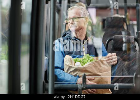 Uomo anziano che guarda la finestra in autobus mentre si viaggia con i mezzi pubblici in città e che tiene una borsa di carta con generi alimentari, spazio copia Foto Stock