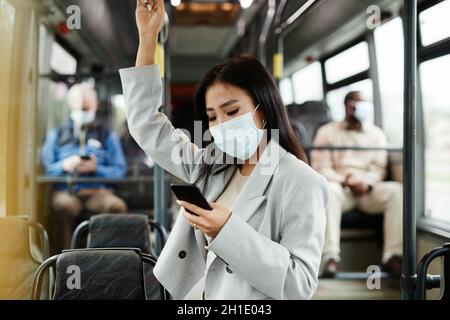 Ritratto della giovane donna asiatica con maschera in autobus e smartphone mentre si è in viaggio in città Foto Stock