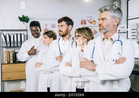 Medico maschile africano americano in fila con colleghi multirazziali, sorridendo e mostrando il pollice in su sulla macchina fotografica. Team di specialisti medici in posa presso il moderno gabinetto. Foto Stock