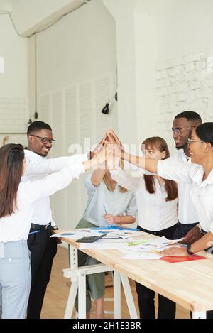 Un gruppo multietnico di dipendenti celebra insieme il successo aziendale. Eccitati i team di affari africani e caucasici che danno cinque alti in un incontro d'ufficio Foto Stock
