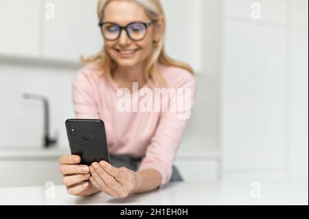 Sorridente ragazza bionda affascinante trascorre il tempo libero con uno smartphone a casa, controllare le e-mail, shopping online, scorrimento news feed, sorridente mezza età sesso femminile web surf, fuoco selettivo al telefono Foto Stock